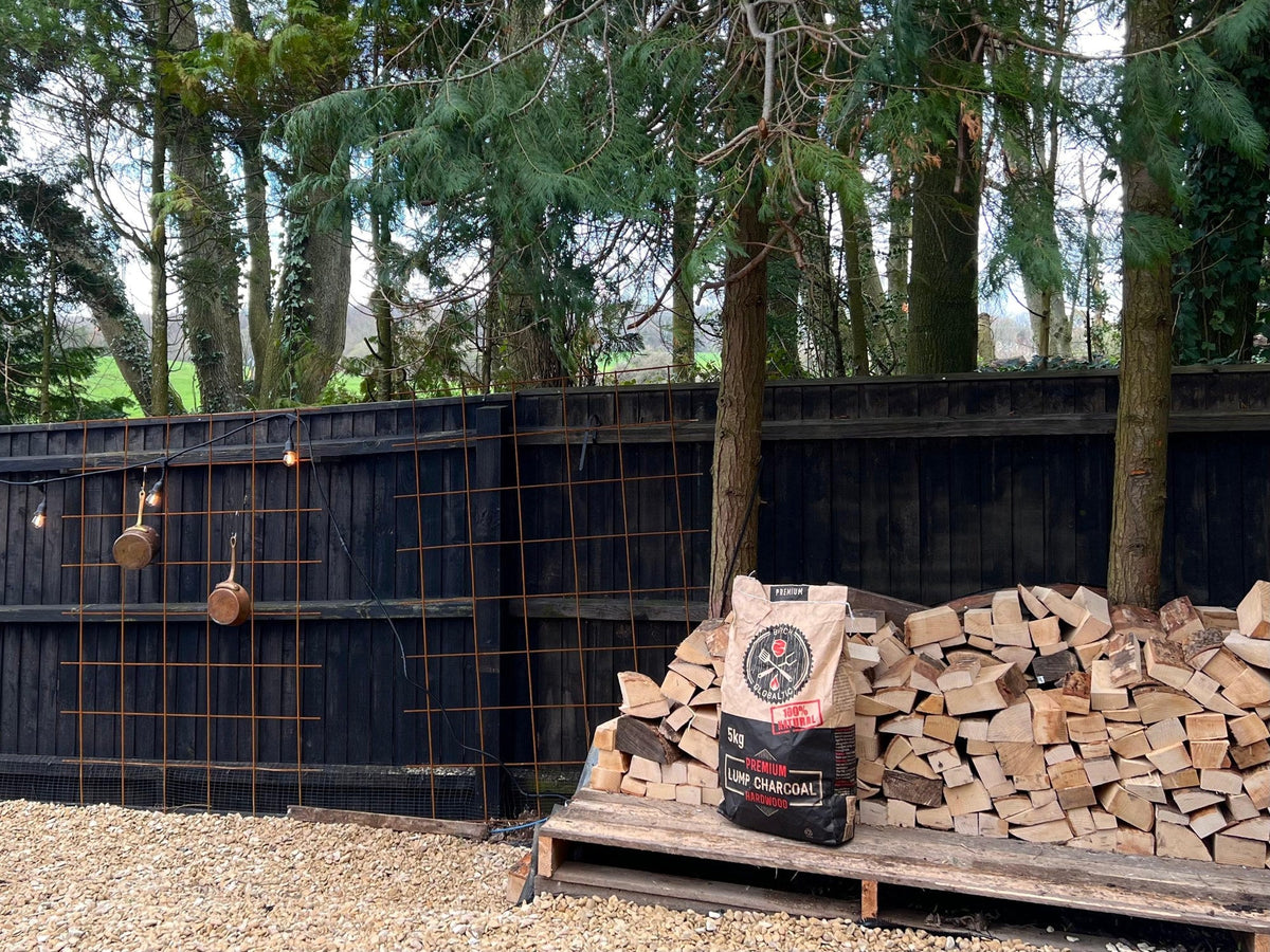 A bag of Globaltic premium lumpwood charcoal on a pallet beside stacked hardwood logs in an outdoor cooking area